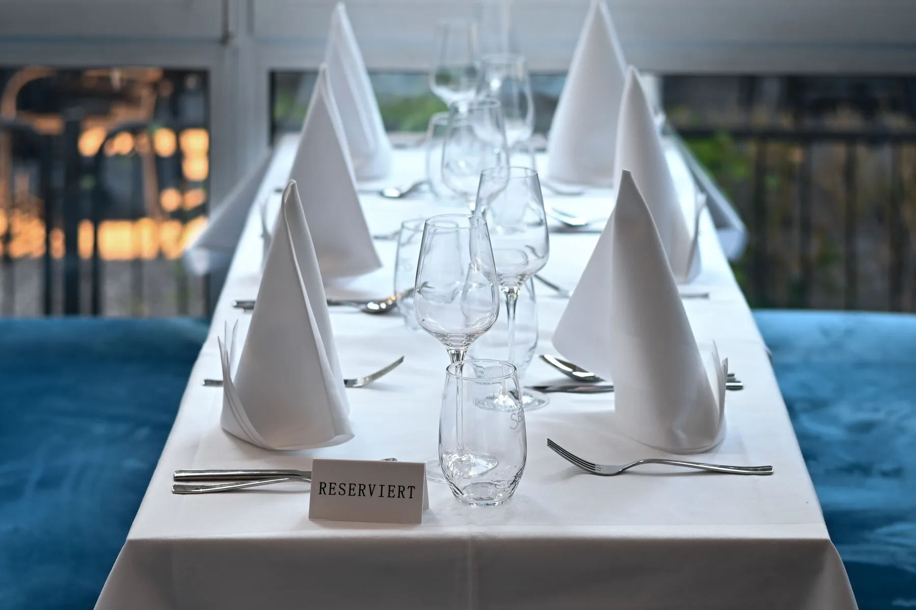 Eine lange, weiß eingedeckte Tafel zwischen zwei blauen Samtbänken, mit Serviettenfächern, Gläsern und einem Kärtchen RESERVIERT